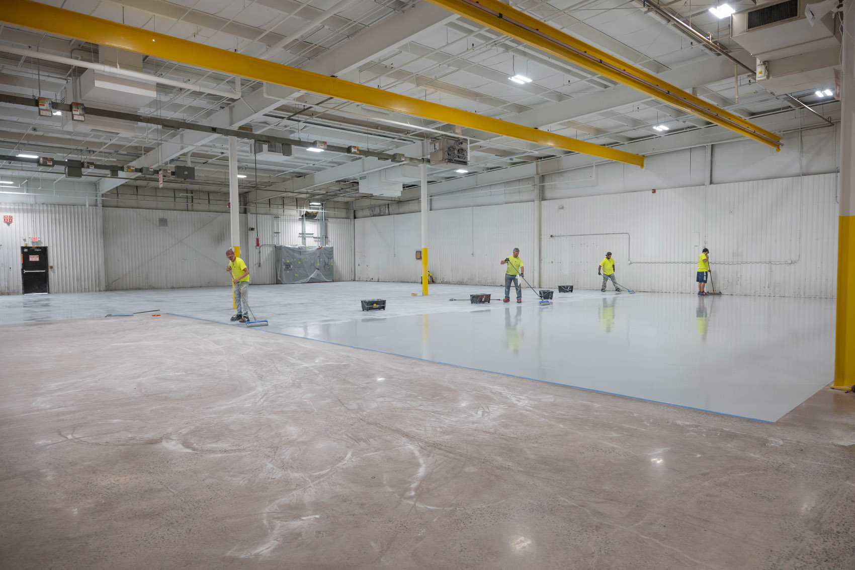 Four workers in yellow shirts apply a light-colored coating to the floor of a large, bright industrial warehouse with high ceilings and exposed pipes. The foreground shows unfinished concrete.