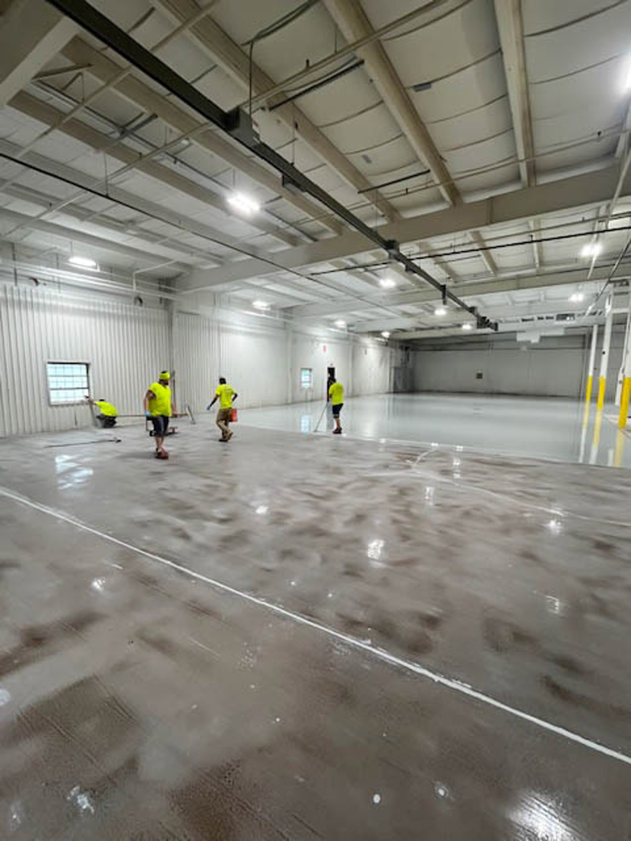 Four workers in yellow shirts are cleaning or treating the floor of a large, empty industrial warehouse with high ceilings and overhead lighting. The floor appears partially wet or freshly coated.