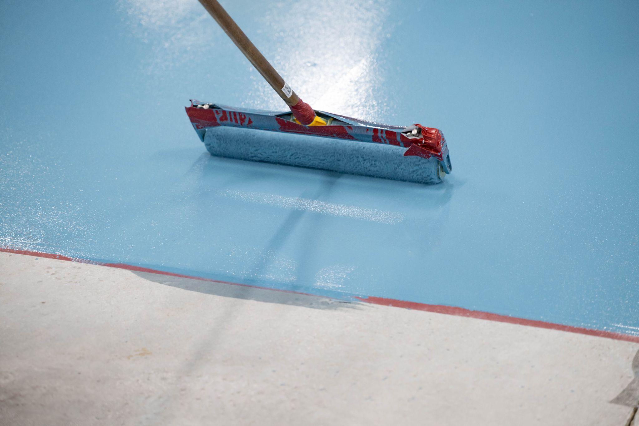 A paint roller with a long handle is applying light blue paint to a concrete floor, covering the surface smoothly. The roller and edge of the painted area are clearly visible.