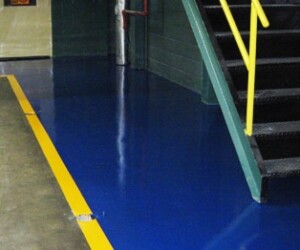 A blue industrial floor coating with a yellow safety line runs beside a green metal staircase with a yellow handrail in an industrial setting. A blue industrial floor coating with a yellow safety line runs beside a green metal staircase with a yellow handrail in an industrial setting.