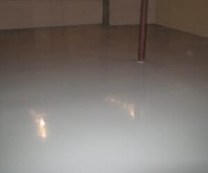 A smooth, shiny, light gray concrete floor in an empty indoor space with a single vertical brown metal support pole. Reflections of ceiling lights highlight the polished industrial floor coating on this surface. A smooth, shiny, light gray concrete floor in an empty indoor space with a single vertical brown metal support pole. Reflections of ceiling lights highlight the polished industrial floor coating on this surface.