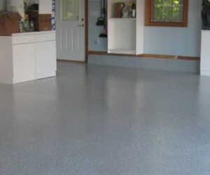 A clean, empty garage with a smooth, anti-slip epoxy floor in speckled gray, white cabinets, stacked boxes, and shelves with various items along the walls. A door and window provide natural light. A clean, empty garage with a smooth, anti-slip epoxy floor in speckled gray, white cabinets, stacked boxes, and shelves with various items along the walls. A door and window provide natural light.