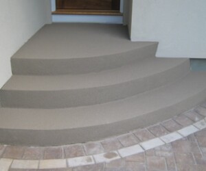 Four wide, curved concrete steps lead up to a doorway, bordered by a light-colored wall and a section of tan brick paving in front featuring durable anti-slip epoxy flooring for added safety. Four wide, curved concrete steps lead up to a doorway, bordered by a light-colored wall and a section of tan brick paving in front featuring durable anti-slip epoxy flooring for added safety.