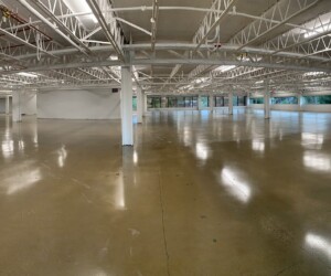 A spacious, empty warehouse with anti-slip epoxy flooring, white support beams, and large windows along the walls allowing natural light to stream in. The ceiling is exposed, revealing pipes and ducts. A spacious, empty warehouse with anti-slip epoxy flooring, white support beams, and large windows along the walls allowing natural light to stream in. The ceiling is exposed, revealing pipes and ducts.