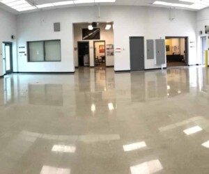 A wide, empty room with shiny, polished concrete floors featuring industrial floor coating, white walls, and a high ceiling with fluorescent lights. Several doors and windows line the far wall, and there is a large rolling garage door on the right. A wide, empty room with shiny, polished concrete floors featuring industrial floor coating, white walls, and a high ceiling with fluorescent lights. Several doors and windows line the far wall, and there is a large rolling garage door on the right.