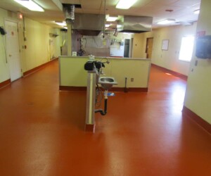 A commercial kitchen with USDA approved anti-slip epoxy flooring in orange-red, cream-colored walls, stainless steel appliances, and a central handwashing sink. Overhead vents and lighting fixtures highlight the clean, organized space. A commercial kitchen with USDA approved anti-slip epoxy flooring in orange-red, cream-colored walls, stainless steel appliances, and a central handwashing sink. Overhead vents and lighting fixtures highlight the clean, organized space.