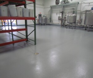 Industrial facility with a smooth, shiny, gray anti-slip epoxy floor, red metal shelving on the left, plastic buckets, and stainless steel tanks and equipment along the back wall. Ideal for spaces needing durable commercial kitchen flooring. Industrial facility with a smooth, shiny, gray anti-slip epoxy floor, red metal shelving on the left, plastic buckets, and stainless steel tanks and equipment along the back wall. Ideal for spaces needing durable commercial kitchen flooring.
