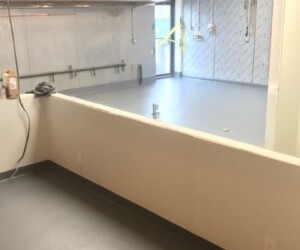 A clean, empty industrial room with white walls, exposed metal fixtures, and gray anti-slip epoxy flooring. A half-wall sits in the foreground, while a door marked with yellow caution tape stands in the background. A clean, empty industrial room with white walls, exposed metal fixtures, and gray anti-slip epoxy flooring. A half-wall sits in the foreground, while a door marked with yellow caution tape stands in the background.