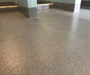 A smooth, speckled gray anti-slip epoxy floor with curved edges and coved base rises up to blue-tiled walls and steps, creating a seamless, clean surface in an indoor setting. A smooth, speckled gray anti-slip epoxy floor with curved edges and coved base rises up to blue-tiled walls and steps, creating a seamless, clean surface in an indoor setting.