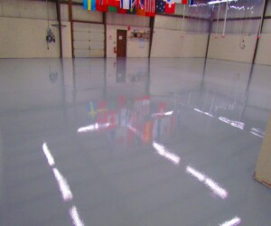 A large, empty warehouse with a shiny, polished epoxy flooring Connecticut reflecting ceiling lights and hanging international flags. Beige walls and a closed overhead door are visible in the background. A large, empty warehouse with a shiny, polished epoxy flooring Connecticut reflecting ceiling lights and hanging international flags. Beige walls and a closed overhead door are visible in the background.