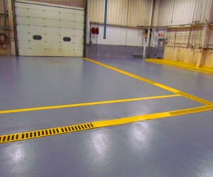 A clean, empty warehouse interior with smooth, shiny gray epoxy flooring Connecticut, yellow lines and grates, a large garage door, wall-mounted hoses, and industrial walls. A clean, empty warehouse interior with smooth, shiny gray epoxy flooring Connecticut, yellow lines and grates, a large garage door, wall-mounted hoses, and industrial walls.