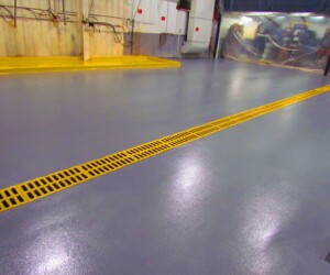 A clean, shiny industrial floor with a bright yellow grated trench drain running through the center showcases durable industrial floor coating. The walls have utility panels and piping, with some plastic sheeting in the background. A clean, shiny industrial floor with a bright yellow grated trench drain running through the center showcases durable industrial floor coating. The walls have utility panels and piping, with some plastic sheeting in the background.