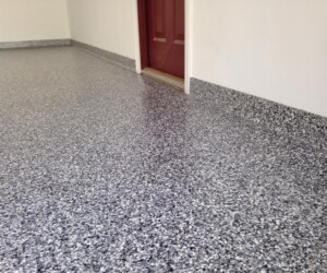 A clean garage floor with a gray and black speckled USDA approved epoxy flooring, matching baseboards, cream-colored walls, and a red door in the background. A clean garage floor with a gray and black speckled USDA approved epoxy flooring, matching baseboards, cream-colored walls, and a red door in the background.