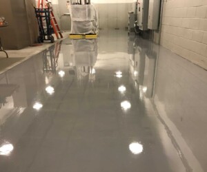 A clean, polished, and shiny gray anti-slip epoxy floor reflects ceiling lights in an industrial hallway with equipment, a red ladder, and covered objects along the walls. A clean, polished, and shiny gray anti-slip epoxy floor reflects ceiling lights in an industrial hallway with equipment, a red ladder, and covered objects along the walls.