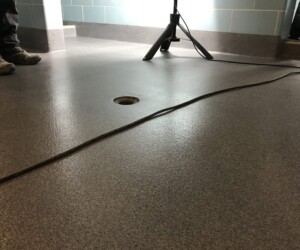 A textured gray industrial floor coating with a central drain, a cord running across it, and two people standing nearby; a tripod or light stand is also visible in the background. A textured gray industrial floor coating with a central drain, a cord running across it, and two people standing nearby; a tripod or light stand is also visible in the background.