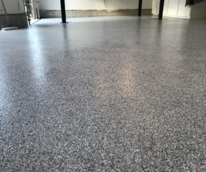 Close-up view of a smooth, speckled gray industrial floor coating on a garage floor, with the bottom part of the garage walls and some equipment visible in the background. Close-up view of a smooth, speckled gray industrial floor coating on a garage floor, with the bottom part of the garage walls and some equipment visible in the background.