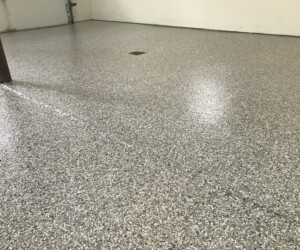 A clean, empty garage with a smooth, speckled gray epoxy-coated floor—an example of quality epoxy flooring Connecticut—plus a square drain in the center, a support pole to the left, and off-white walls in the background. A clean, empty garage with a smooth, speckled gray epoxy-coated floor—an example of quality epoxy flooring Connecticut—plus a square drain in the center, a support pole to the left, and off-white walls in the background.