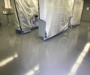 A clean industrial room with a smooth, shiny gray resinous flooring CT. Large equipment is covered with white plastic sheeting and blue tape, suggesting protection during maintenance or painting work. A clean industrial room with a smooth, shiny gray resinous flooring CT. Large equipment is covered with white plastic sheeting and blue tape, suggesting protection during maintenance or painting work.