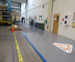 A factory floor features a safety-marked walkway, yellow caution tape, cones, and wall-mounted safety boards. An orange and white “Stay Safe” sign sits on the clean gray anti-slip epoxy floor near the wall, enhancing workplace safety. A factory floor features a safety-marked walkway, yellow caution tape, cones, and wall-mounted safety boards. An orange and white “Stay Safe” sign sits on the clean gray anti-slip epoxy floor near the wall, enhancing workplace safety.