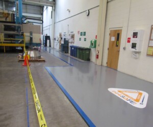 A factory floor features a safety-marked walkway, yellow caution tape, cones, and wall-mounted safety boards. An orange and white “Stay Safe” sign sits on the clean gray anti-slip epoxy floor near the wall, enhancing workplace safety. A factory floor features a safety-marked walkway, yellow caution tape, cones, and wall-mounted safety boards. An orange and white “Stay Safe” sign sits on the clean gray anti-slip epoxy floor near the wall, enhancing workplace safety.