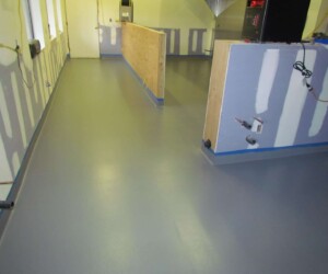 A room under construction with unfinished walls, exposed outlets and pipes, partial partitions, and a smooth, newly-installed gray anti-slip epoxy floor. Natural light enters through several windows on the left side. A room under construction with unfinished walls, exposed outlets and pipes, partial partitions, and a smooth, newly-installed gray anti-slip epoxy floor. Natural light enters through several windows on the left side.