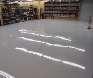 A large room with a shiny, newly finished gray industrial floor coating; shelves filled with bins and equipment line the walls, and caution tape blocks off part of the space. A large room with a shiny, newly finished gray industrial floor coating; shelves filled with bins and equipment line the walls, and caution tape blocks off part of the space.