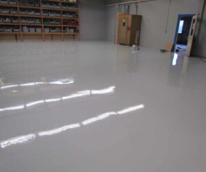 A warehouse with a shiny, freshly coated epoxy flooring Connecticut, shelves along the left wall, and machinery near a wooden door in the back. The polished floor reflects light from overhead fixtures. A warehouse with a shiny, freshly coated epoxy flooring Connecticut, shelves along the left wall, and machinery near a wooden door in the back. The polished floor reflects light from overhead fixtures.