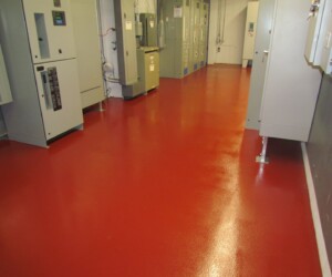 A room with a shiny, red anti-slip epoxy floor, lined with gray electrical control panels and cabinets along both walls. The space is clean and organized, with industrial equipment visible. A room with a shiny, red anti-slip epoxy floor, lined with gray electrical control panels and cabinets along both walls. The space is clean and organized, with industrial equipment visible.