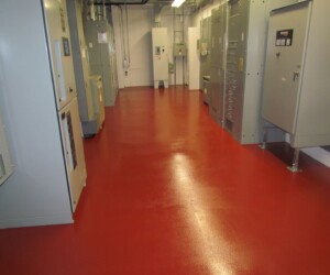 A room with a shiny red industrial floor coating, lined with large grey electrical panels and control cabinets along the walls, with exposed pipes and wiring on the ceiling. A room with a shiny red industrial floor coating, lined with large grey electrical panels and control cabinets along the walls, with exposed pipes and wiring on the ceiling.