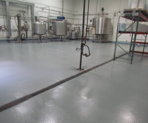 A clean industrial facility with a smooth, gray epoxy flooring Connecticut finish, stainless steel tanks, metal shelving, exposed pipes, and a floor drain; the environment appears bright and well-maintained. A clean industrial facility with a smooth, gray epoxy flooring Connecticut finish, stainless steel tanks, metal shelving, exposed pipes, and a floor drain; the environment appears bright and well-maintained.