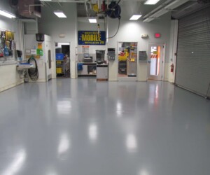 A clean, empty auto workshop with an anti-slip epoxy floor, a garage door on the right, tools hanging on the left wall, and three rooms visible in the back with desks and equipment inside. A clean, empty auto workshop with an anti-slip epoxy floor, a garage door on the right, tools hanging on the left wall, and three rooms visible in the back with desks and equipment inside.