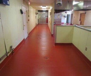 A clean, empty hallway in a commercial kitchen showcases red commercial kitchen flooring, cream-colored walls, overhead lighting, and exposed pipes and ducts. Various doors and kitchen equipment line the industrial corridor. A clean, empty hallway in a commercial kitchen showcases red commercial kitchen flooring, cream-colored walls, overhead lighting, and exposed pipes and ducts. Various doors and kitchen equipment line the industrial corridor.