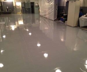 A smooth, shiny gray anti-slip epoxy floor in an industrial room with equipment, cabinets, and plastic sheeting along the walls; overhead lights reflect on the polished surface. A smooth, shiny gray anti-slip epoxy floor in an industrial room with equipment, cabinets, and plastic sheeting along the walls; overhead lights reflect on the polished surface.