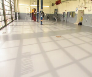 A clean, empty auto service garage with a shiny, resinous flooring CT finish, tall windows casting grid-like shadows, car lifts, wall-mounted equipment, and stairs leading to an upper area in the background. A clean, empty auto service garage with a shiny, resinous flooring CT finish, tall windows casting grid-like shadows, car lifts, wall-mounted equipment, and stairs leading to an upper area in the background.