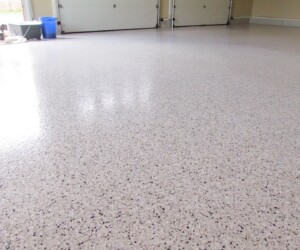 A close-up view of a clean, smooth, speckled epoxy-coated garage floor. The empty garage has light walls, a closed double garage door, and some storage bins in the background—showcasing premium epoxy flooring Connecticut trusts. A close-up view of a clean, smooth, speckled epoxy-coated garage floor. The empty garage has light walls, a closed double garage door, and some storage bins in the background—showcasing premium epoxy flooring Connecticut trusts.