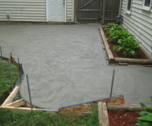 Freshly poured and smoothed concrete patio next to a house with garden beds and green plants along the side, featuring wooden edging and forms—an ideal surface for adding anti-slip epoxy floor protection. Freshly poured and smoothed concrete patio next to a house with garden beds and green plants along the side, featuring wooden edging and forms—an ideal surface for adding anti-slip epoxy floor protection.