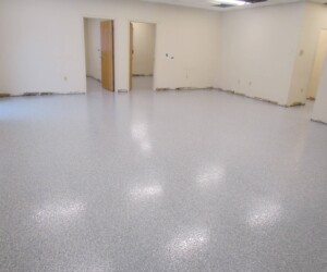 A large, empty room with white walls, light-colored speckled industrial floor coating, and three doorways leading to adjoining rooms. The baseboards are missing, exposing the lower wall edges. A large, empty room with white walls, light-colored speckled industrial floor coating, and three doorways leading to adjoining rooms. The baseboards are missing, exposing the lower wall edges.