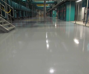 A shiny, light gray USDA approved flooring system coats the industrial floor in a large room with metal stairs on the left, blue machinery and tanks along the walls, and electrical panels on the right. A shiny, light gray USDA approved flooring system coats the industrial floor in a large room with metal stairs on the left, blue machinery and tanks along the walls, and electrical panels on the right.