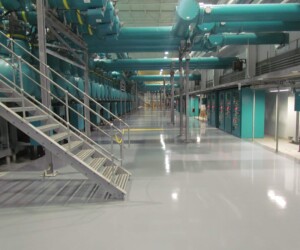 A clean, shiny industrial facility with large teal pipes overhead, control panels on the right, and a metal staircase on the left. The polished, reflective look comes from durable industrial floor coating, and the space is brightly lit. A clean, shiny industrial facility with large teal pipes overhead, control panels on the right, and a metal staircase on the left. The polished, reflective look comes from durable industrial floor coating, and the space is brightly lit.