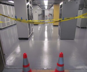 A hallway in a server room is blocked by yellow caution tape and two orange traffic cones, indicating restricted access. The industrial floor coating reflects the surrounding server cabinets and bright overhead lights. A hallway in a server room is blocked by yellow caution tape and two orange traffic cones, indicating restricted access. The industrial floor coating reflects the surrounding server cabinets and bright overhead lights.