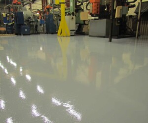 A shiny, freshly coated USDA approved flooring reflects light and nearby machinery in a large manufacturing facility. Various blue and gray machines are visible in the background. A shiny, freshly coated USDA approved flooring reflects light and nearby machinery in a large manufacturing facility. Various blue and gray machines are visible in the background.