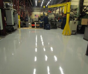 A clean, shiny factory floor features an anti-slip epoxy floor with various industrial machines, barrels, and equipment along the sides. Overhead lights reflect off the polished surface. An American flag hangs on the back wall. A clean, shiny factory floor features an anti-slip epoxy floor with various industrial machines, barrels, and equipment along the sides. Overhead lights reflect off the polished surface. An American flag hangs on the back wall.