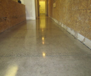A long hallway with polished concrete floors, unfinished chipboard walls, and exposed base areas features industrial floor coating. The hallway leads to a brightly lit doorway at the end. A long hallway with polished concrete floors, unfinished chipboard walls, and exposed base areas features industrial floor coating. The hallway leads to a brightly lit doorway at the end.