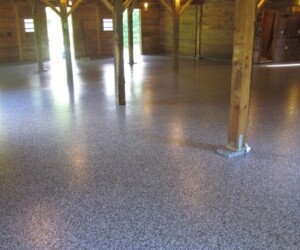 Interior of a wooden barn or large shed with exposed support beams, multiple small windows, and a shiny, gray-speckled USDA approved flooring. Sunlight is visible through the windows in the background. Interior of a wooden barn or large shed with exposed support beams, multiple small windows, and a shiny, gray-speckled USDA approved flooring. Sunlight is visible through the windows in the background.