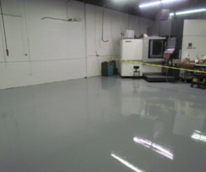 A clean, shiny, gray anti-slip epoxy floor in an industrial workspace, with caution tape cordoning off an area near a large machine and various equipment against white walls under fluorescent lighting. A clean, shiny, gray anti-slip epoxy floor in an industrial workspace, with caution tape cordoning off an area near a large machine and various equipment against white walls under fluorescent lighting.