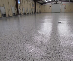 A smooth, shiny industrial floor coating covers the concrete inside a spacious building with beige walls, radiators, and a large garage door at the far end. A smooth, shiny industrial floor coating covers the concrete inside a spacious building with beige walls, radiators, and a large garage door at the far end.