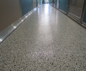 A polished terrazzo hallway floor with a speckled pattern, bordered by metal and glass walls, features industrial floor coating and stretches toward a distant bright area at the end. A polished terrazzo hallway floor with a speckled pattern, bordered by metal and glass walls, features industrial floor coating and stretches toward a distant bright area at the end.
