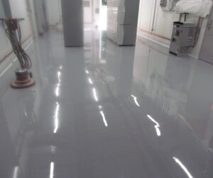 A hallway with a shiny, newly coated gray epoxy flooring Connecticut finish, cleaning equipment, and some covered machinery along the walls under fluorescent ceiling lights. A hallway with a shiny, newly coated gray epoxy flooring Connecticut finish, cleaning equipment, and some covered machinery along the walls under fluorescent ceiling lights.