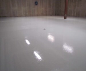 A large indoor space with smooth, shiny, freshly coated anti-slip epoxy flooring reflects overhead lights. The room has unfinished concrete walls, a single red support column, and a small floor drain in the center. A large indoor space with smooth, shiny, freshly coated anti-slip epoxy flooring reflects overhead lights. The room has unfinished concrete walls, a single red support column, and a small floor drain in the center.