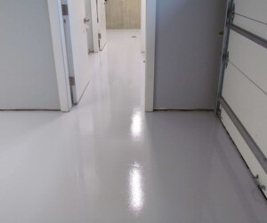 A clean, shiny, light gray epoxy-coated garage floor—similar to USDA approved flooring used in commercial kitchen flooring—leads to several white doors and a partially open garage door. The surface reflects light, creating a glossy finish. A clean, shiny, light gray epoxy-coated garage floor—similar to USDA approved flooring used in commercial kitchen flooring—leads to several white doors and a partially open garage door. The surface reflects light, creating a glossy finish.